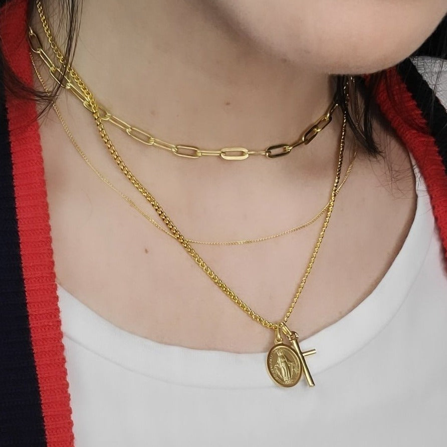 Solid Yellow Gold Cross Charm on a 3mm Wide Curb Chain Layered with other Necklaces and Pendants Worn by a Woman